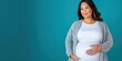© SerPak - Pregnant hispanic female in casual attire holding belly against blue background.