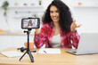 © sofiko14 - Young adult African woman recording video blog using smartphone on tripod at home office setting. She is wearing a plaid shirt while presenting her content through the camera in a casual setup.
