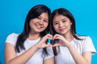 © Daniel - Two Asian teen girls smiling and forming a heart shape with their hands, standing close together in white tops and plaid skirts, against a solid light blue background