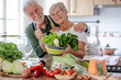 © luciano - Attractive senior couple working together in home kitchen preparing vegetables. Caucasian elderly smiling people retirees enjoying healthy eating looking at camera
