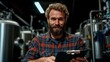 ©  Shomixer - A cheerful man with a beard smiles genuinely while holding a clipboard in a well-lit brewery, representing enjoyment and involvement in the craft of brewing.