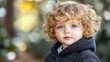 © JO BLA CO - A young child with curly blonde hair and captivating blue eyes stands outdoors, gazing curiously at the surroundings. The background is softly blurred, hinting at a serene natural environment