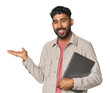 © Asier - Hispanic male holding laptop in studio showing a copy space on a palm and holding another hand on waist.