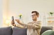 © Studio Romantic - Serious man in glasses sitting on couch at home, holding smartphone at arm length, trying to discern. Young guy in eyewear squinting at screen, experiencing vision problems or blurred eyesight.