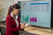 © Wasana - A woman in a red jacket is writing on a clipboard in front of a large monitor