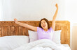 © Prostock-studio - Good morning. Happy asian woman yawning waking up sitting in cozy bed at home on weekend.