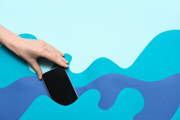Wall Mural - female hand hiding mobile phone in paper waves on blue background