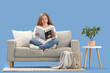 © Pixel-Shot - Beautiful young woman reading magazine on stylish grey sofa against blue background