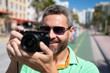 © Volodymyr - A traveler with camera taking photo on streets of Miami. A man traveler enjoys his summer vacation in a tropical city. A traveler tourist with suitcase on summer city. Man traveler walk on city.