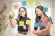 © MINAE - Two Asian businesswomen collaborating at a glass wall, using vibrant sticky notes to brainstorm ideas and develop a strategic plan for their company's growth and success