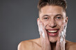 © New Africa - Smiling man washing his face with cleansing foam on grey background. Space for text