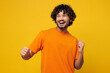 © ViDi Studio - Young smiling fun happy Indian man wear orange t-shirt casual clothes doing winner gesture celebrate clenching fists dancing look aside on area isolated on plain yellow background. Lifestyle concept.