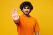 © ViDi Studio - Young sad mad strict serious Indian man he wearing orange t-shirt casual clothes showing stop gesture with palm looking camera isolated on plain yellow background studio portrait. Lifestyle concept.