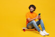 © ViDi Studio - Full body young Indian man he wear orange t-shirt casual clothes sit on skateboard hold use mobile cell phone look aside on area isolated on plain yellow background studio portrait. Lifestyle concept.