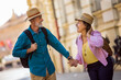 © Mediteraneo - Elderly married couple exploring new city. They are holding hands and wearing hats and backpacks.