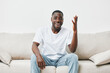 © SHOTPRIME STUDIO - Joyful Black man sitting on a sofa, expressing enthusiasm and engagement, wearing a casual white t shirt and light blue jeans in a minimalist living room