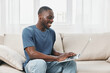 © SHOTPRIME STUDIO - Young Black man smiling while using a laptop at home, dressed casually in a blue t shirt and jeans, showcasing a relaxed and happy lifestyle