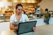 © DusanJelicic - Female baker working on a laptop, managing online orders and inventory for her bakery, while customers happily purchase fresh bread in the bustling background