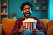 © miss irine - Excited indian man with beard in glasses, red checkered shirt eats popcorn, watching movie on sofa at home. Guy enjoys tv show, entertainment online content, laughs hard. Weekend relax lifestyle.