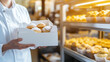 © Curioso.Photography - A baker in a white uniform holding a box of freshly baked pastries in a bakery filled with golden bread and sweets.
