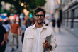 © BullRun - young man with relaxed smile stands confidently on city street, holding tablet. His casual yet professional outfit and wireless earbuds suggest, modern freelancer or student embracing digital work