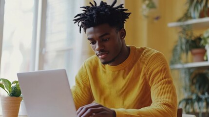 Portrait of positive young black guy in yellow sweater sitting at table, working on laptop, sending emails, office interior. Cheerful african american man freelancer working from home, copy space