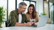 © TimosBlickfang - Happy middle-aged couple using smartphone and laptop together, smiling and enjoying online browsing at home, modern interior with natural light
