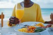 © Iryna - A woman is holding a glass of orange juice and sitting at a table with a plate of food in front of her