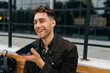 © dikushin - Close-up of cheerful young male recording voice message on smartphone while sitting on bench in city street, enjoying moment of relaxation during workday. Concept of modern successful city lifestyle.