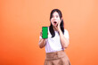 © Ajab - A woman asia wearing a white shirt and brown skirt holding a smart phone device with a green screen of a surprised expression with an open mouth and one hand raised isolated on an orange background
