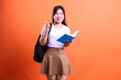 © Ajab - Young woman with a book and a pen standing with a backpack on an orange background ready to study
