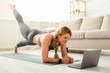 © Prostock-studio - Online sport during quarantine at home. Strong fitness woman in sportswear does exercises on mat on floor and looks at laptop in living room interior, free space