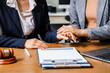 © Phushutter - Two female lawyers in business suits hold hands at a wooden desk, offering comfort and encouragement. A wooden gavel and scales symbolize justice as they discuss corporate law, compliance