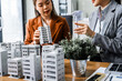 © Phushutter - Two businesswomen sit at a desk in a meeting, discussing property development while building a model of city tower. real estate investments, zoning laws, sustainable energy solutions. net zero