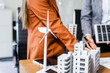 © Phushutter - Two businesswomen sit at a desk in a meeting, discussing property development while building a model of city tower. real estate investments, zoning laws, sustainable energy solutions. net zero