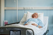© Seventyfour - Senior patient resting in hospital bed wearing hospital gown and looking contemplative. Electrical outlets and medical equipment visible on wall behind patient