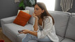 © Krakenimages.com - Young woman watching television in her apartment living room, appearing thoughtful while holding a remote control and sitting on a gray couch with colorful pillows in a cozy home interior.