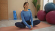 © Krakenimages.com - Woman meditating in gym, sitting cross-legged on mat with eyes closed, exercising in tranquil indoor setting with yoga props and exercise balls in background