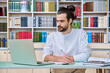 © Valerii Honcharuk - Young male teacher working in library sitting at desk with laptop books