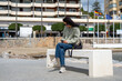 © sutulastock - A young woman with curly dark hair, wearing sunglasses, a green jacket, and jeans, sits on a white bench near the beach, looking at her smartphone with a black shoulder bag beside her