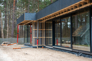 Modern house under construction with black exterior, large glass windows, wooden framing, red support beams, and a visible ventilation system. Tall trees and a wooden fence surround the site.