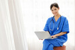 © artitwpd - Smiling Asian healthcare professional in blue scrubs using a laptop, representing compassionate senior healthcare, caregiver support, and telemedicine services for elderly patient well-being.
