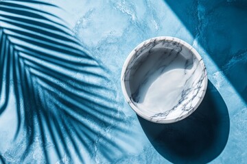 Poster - Beautiful marble bowl casting shadows on a vibrant blue surface with palm leaf reflection during daylight hours