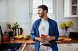 © baranq - Smiling man in kitchen holding cup looking out the window