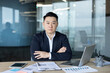 © Tetiana - Portrait of a serious young Asian male businessman, director and owner sitting at a desk in the office, looking confidently at the camera