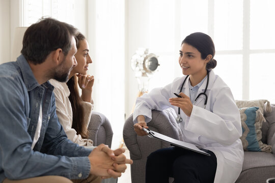 Positive young Indian doctor woman visiting couple at home, writing medical records, filling patient information file, talking to husband and wife, smiling, giving optimistic consultation