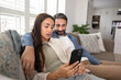 © fizkes - Couple using web service, app on smartphone for Internet communication, shopping, online order, booking, relaxing on couch at home. Husband hugging wife scrolling social media on mobile phone.