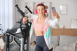 © Pixel-Shot - Sporty young woman with headphones training and mobile phone taking selfie near modern exercising bike at home
