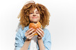 © VICHIZH - Young woman with curly hair joyfully enjoying a delicious burger against a bright white background, showcasing a celebration of healthy eating and happiness