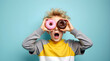 © Gustav - Excited blond boy peering through pink and chocolate donuts with a surprised expression against a light blue backdrop
